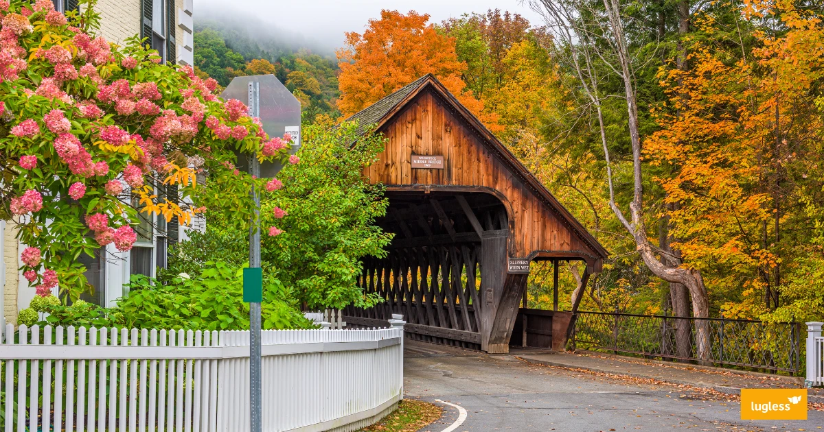 A wooden covered bridge framed by red, orange, and gold fall foliage beside a white picket fence on a quiet small-town street