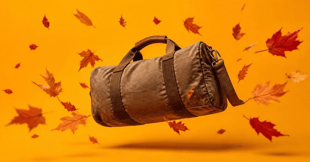A tan canvas duffel bag with brown leather straps floating against a warm amber background, surrounded by falling red and orange autumn leaves
